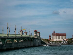 The Bridge of Lions in St. Augustine, Florida, stretching across the Matanzas River with its distinctive green arches and red-roofed towers. The historic downtown skyline, including iconic Spanish-style buildings, is visible in the background on a clear day.